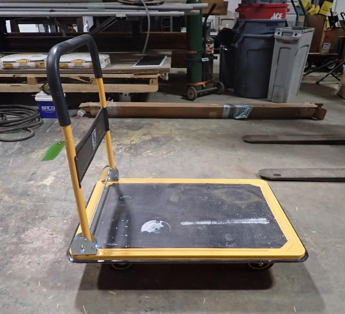 A yellow, four-wheeled flatbed cart with a foldable handle sits on a concrete floor in a workshop or warehouse.