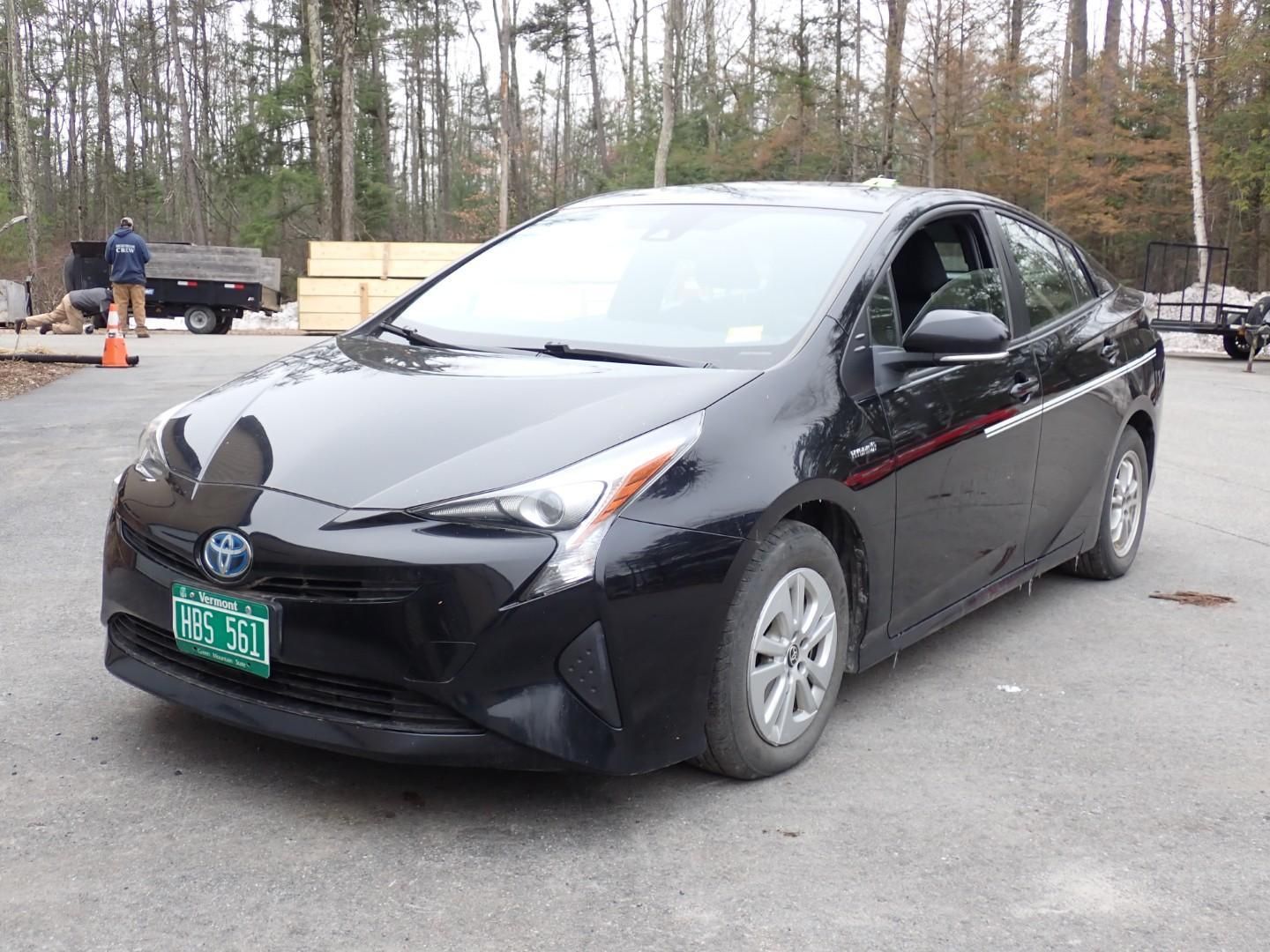A black Toyota Prius parked on a paved lot in front of a wooded area.