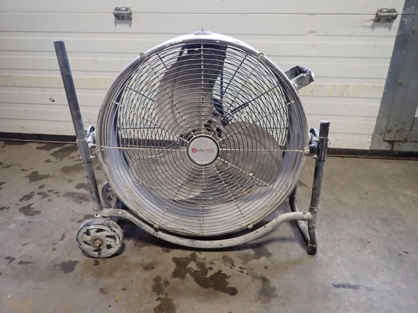 A silver industrial-style floor fan on wheels, standing on a concrete floor in front of a white garage door.