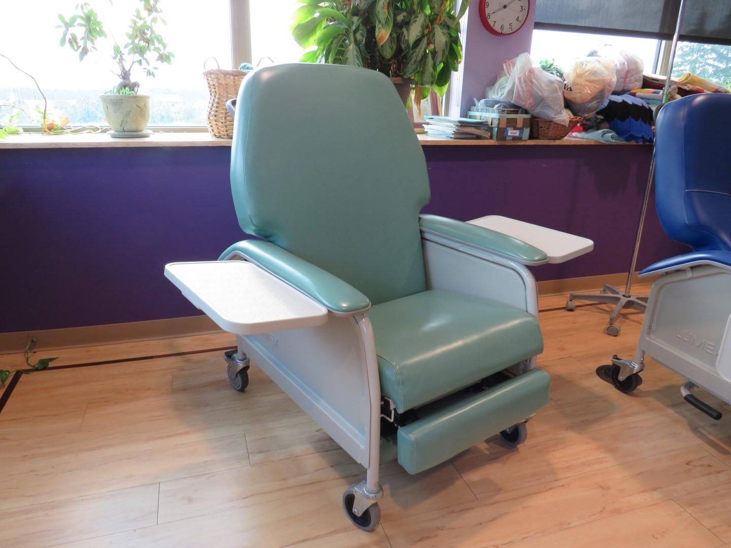 Green medical recliner chair with attached tray tables, wheels, near a window.