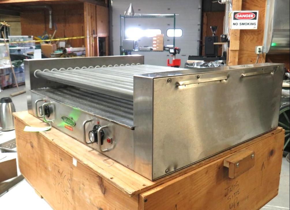 Stainless steel hot dog roller grill on a wooden crate in a workshop.