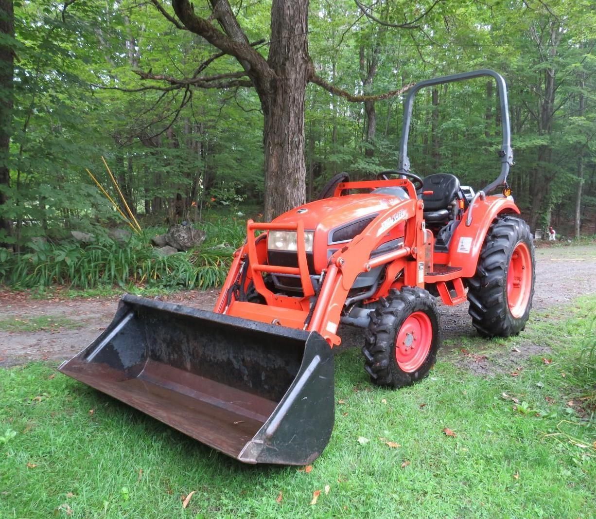Orange tractor with front loader on grass near trees.