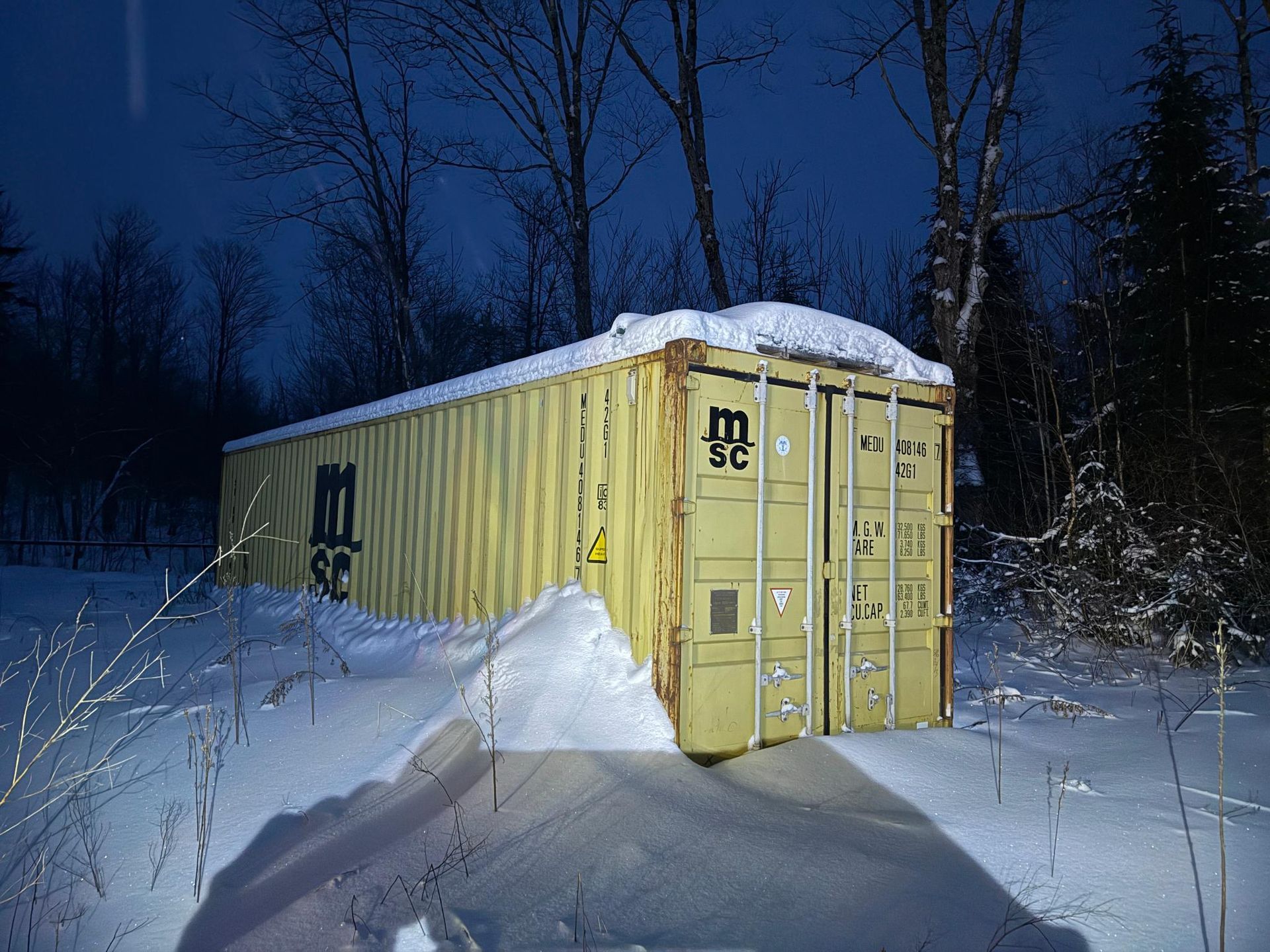 Yellow shipping container covered in snow in a snowy, wooded area at night.