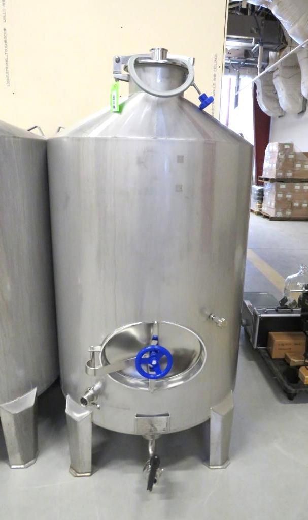 Stainless steel brewing tank with valves and fittings, standing indoors.