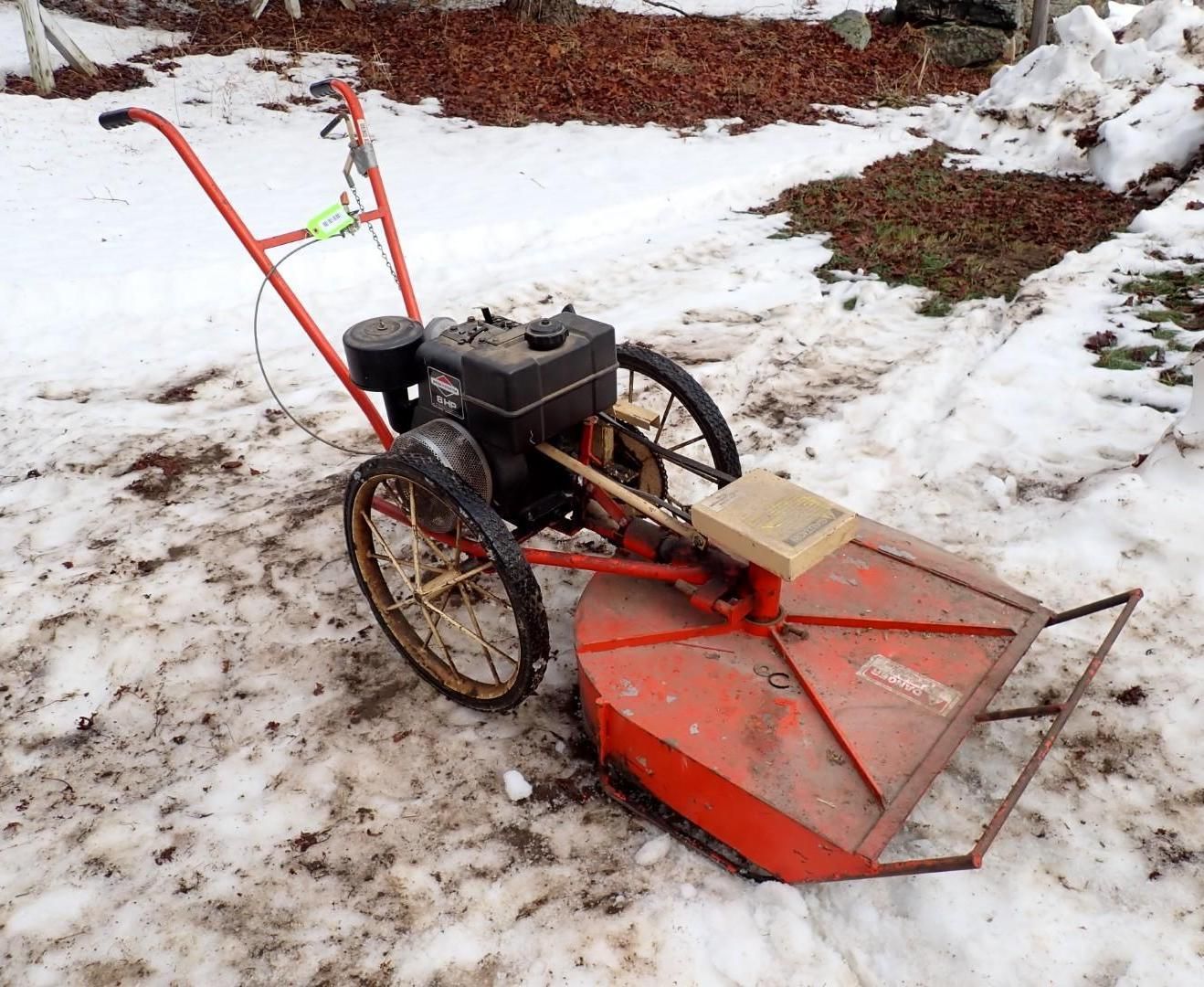 An orange walk-behind brush mower sits on snow-covered ground.