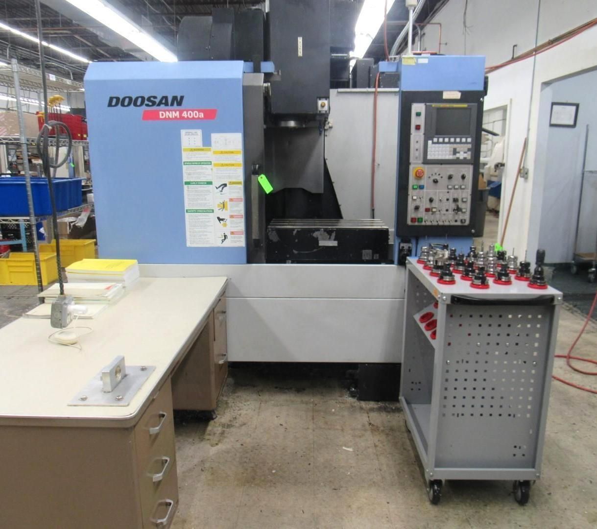 A blue Doosan CNC machine in a workshop with a control panel and tool cart.
