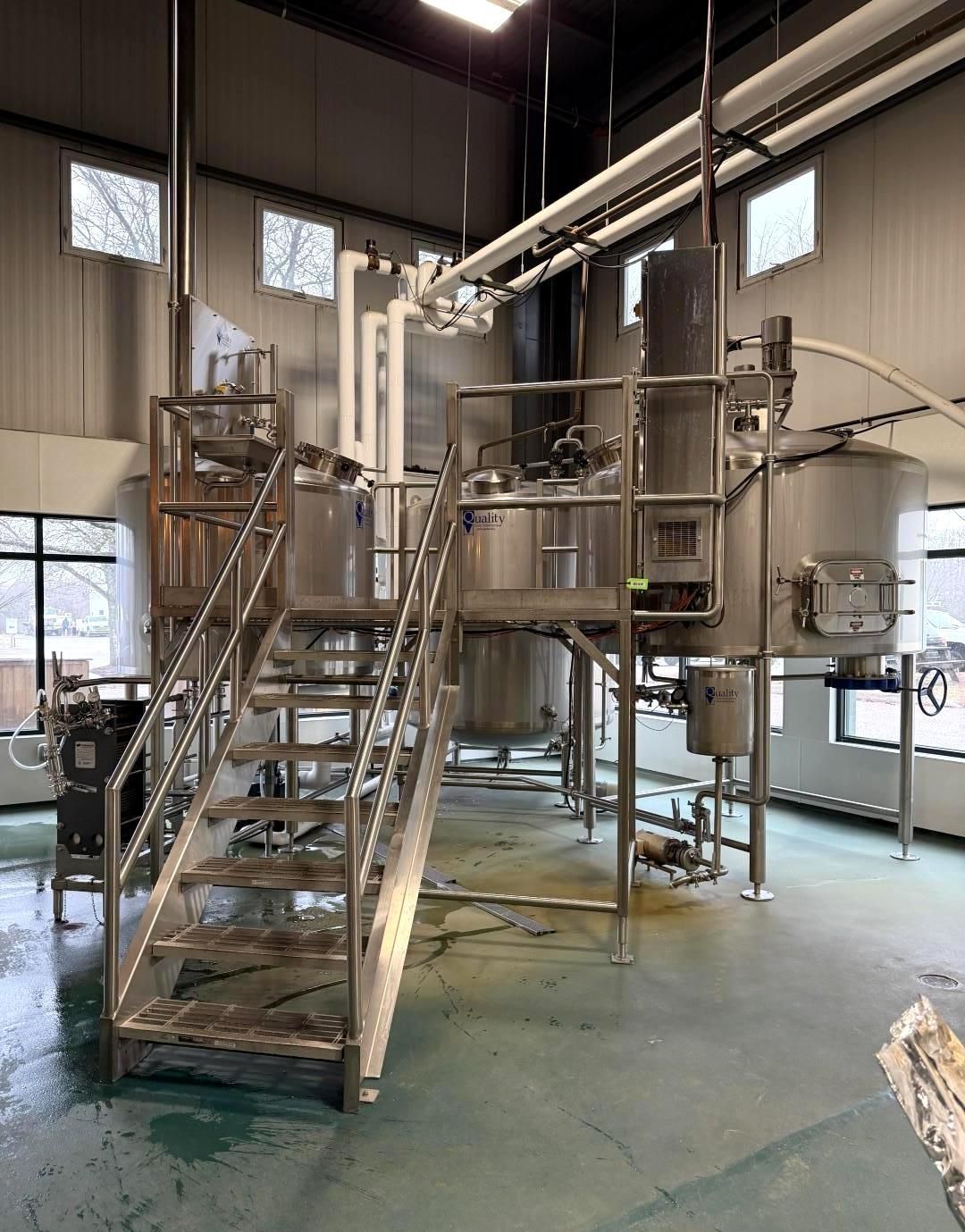 Brewery equipment: stainless steel tanks, stairs, and piping in an industrial setting.