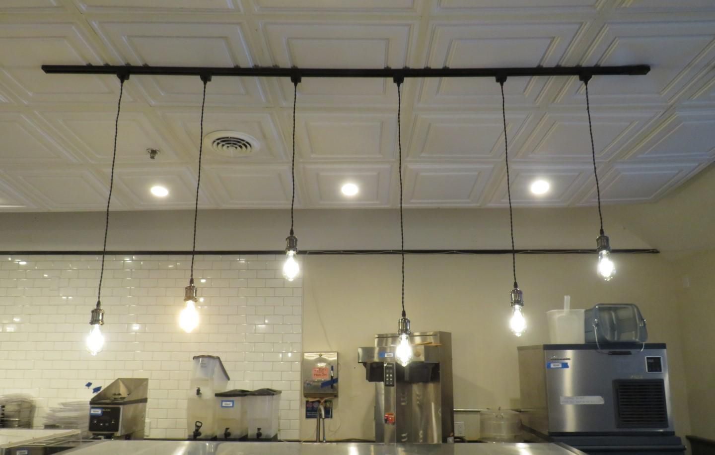 A series of six pendant lights hang above a stainless steel countertop in a commercial kitchen setting.