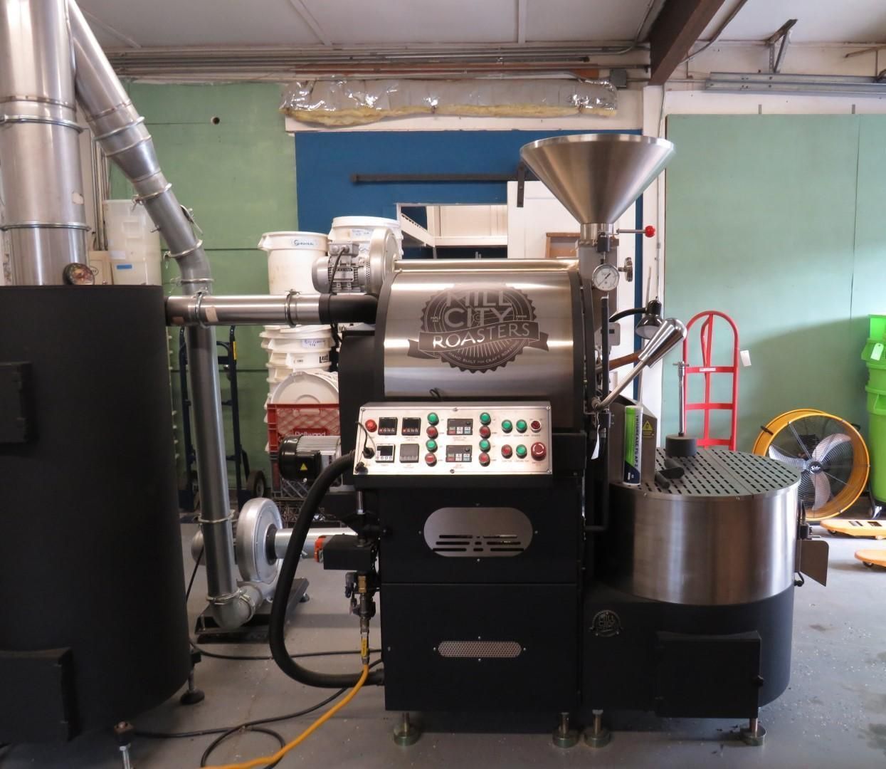A commercial coffee roaster machine in a warehouse setting with a silver hopper and control panel.