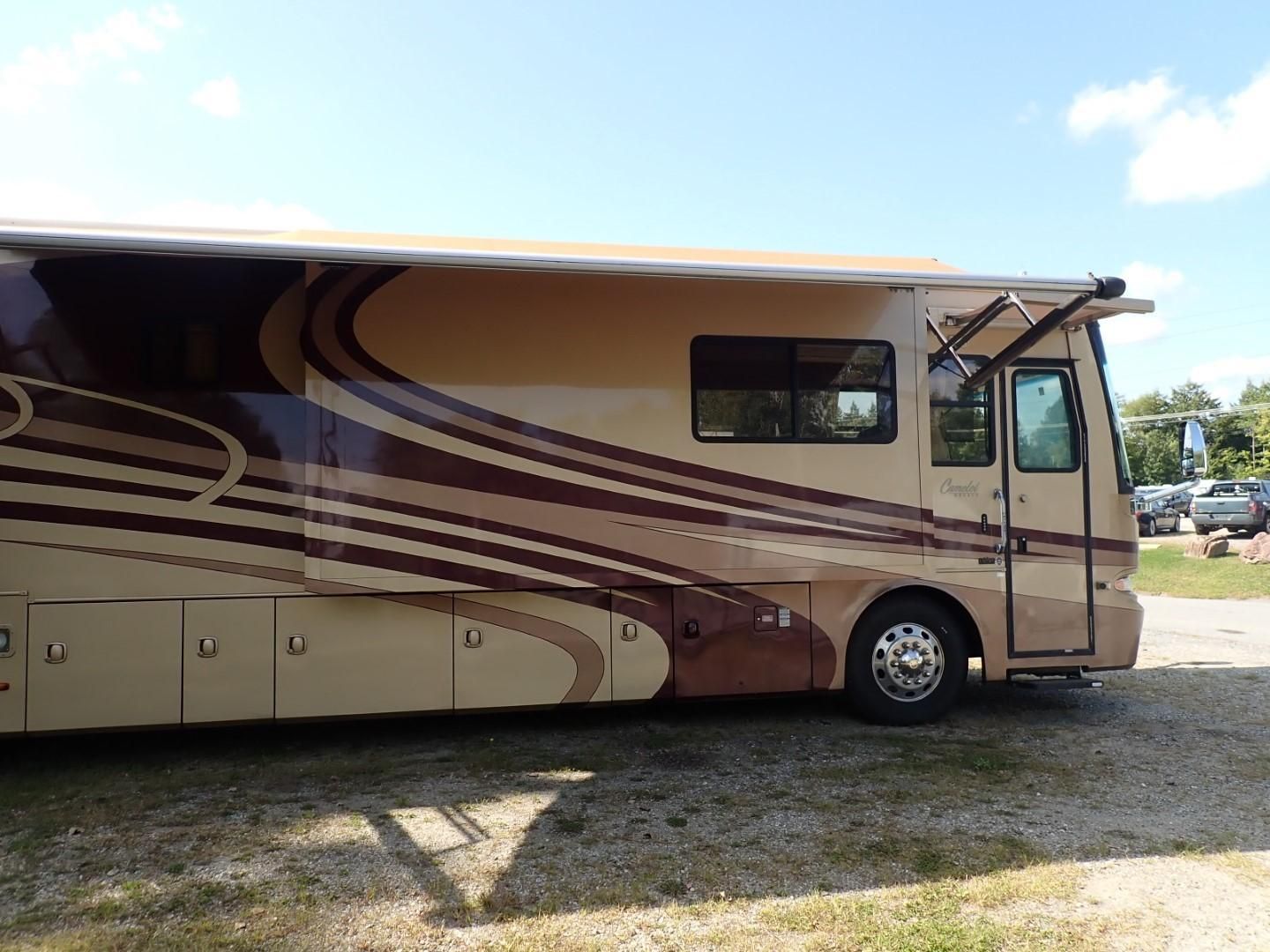 Tan and brown RV parked outdoors with awning extended. Sunny day.