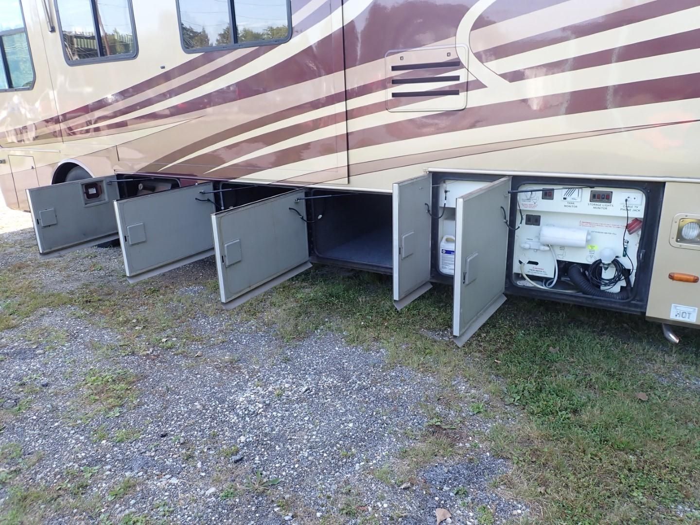 Side of a beige RV with multiple storage compartments open on a gravel surface.
