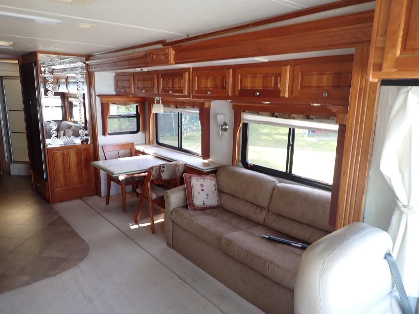 Interior of a recreational vehicle. Light-colored walls, wood cabinets, tan sofa, table with chairs, and windows.