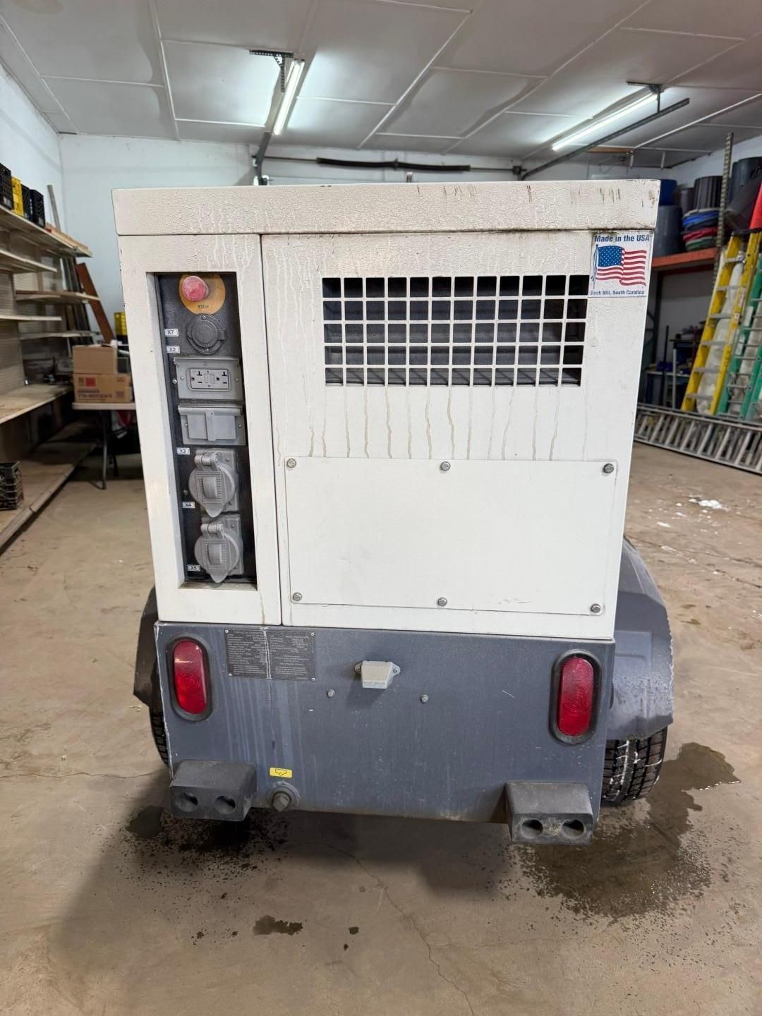 Rear view of a portable generator on wheels, with control panel, grates, and outlets. Grey and white colors.
