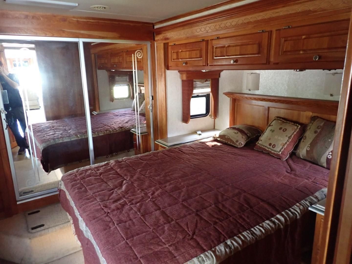 Bedroom interior of a RV with wooden cabinets, a bed with burgundy bedding, and mirrored closet doors.