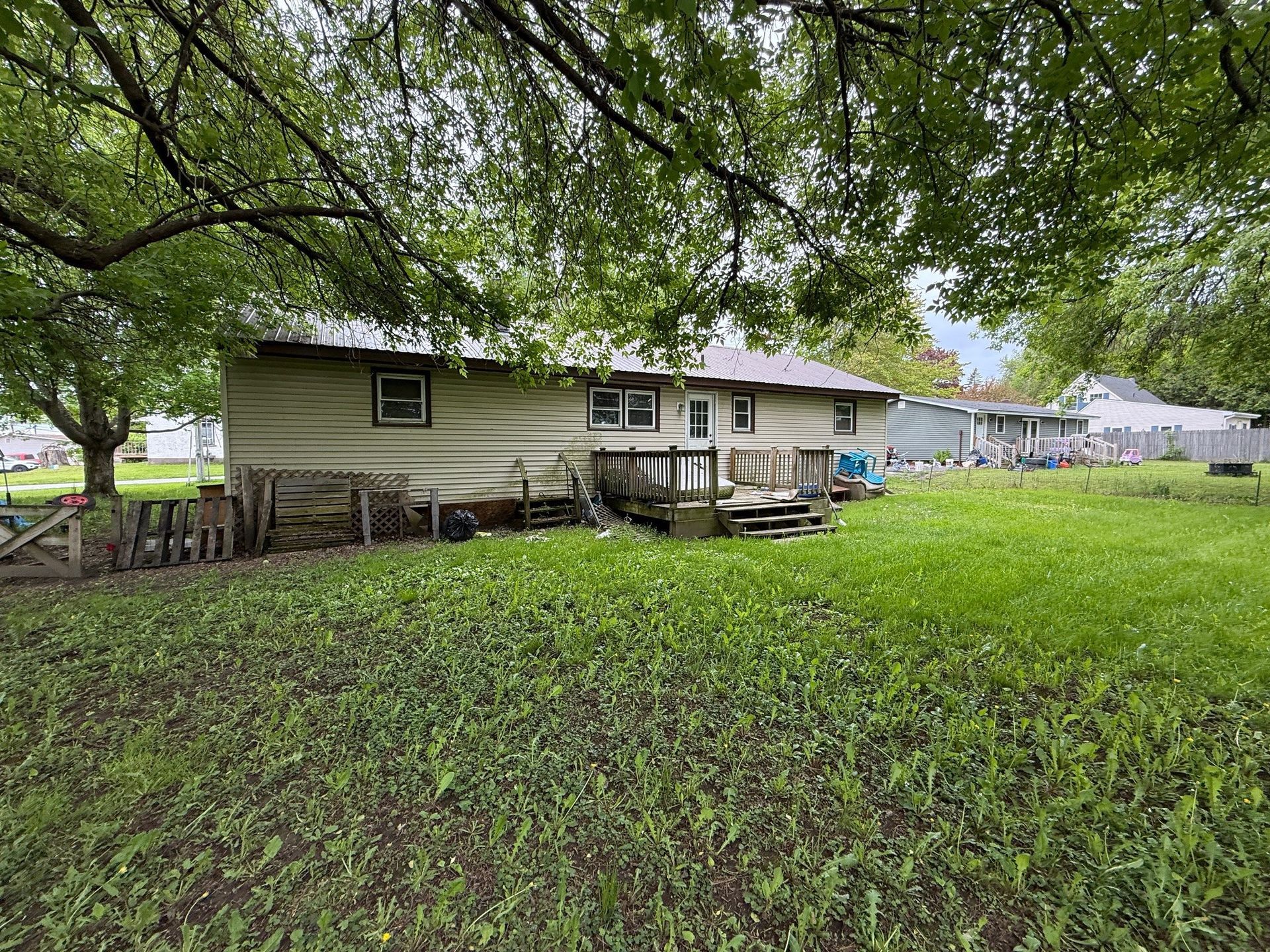 Backyard view of a beige house with a wooden deck, grass, and a tree overhead on a cloudy day.