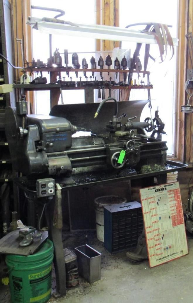 A dark, metal lathe in a workshop, with tools on a shelf behind.