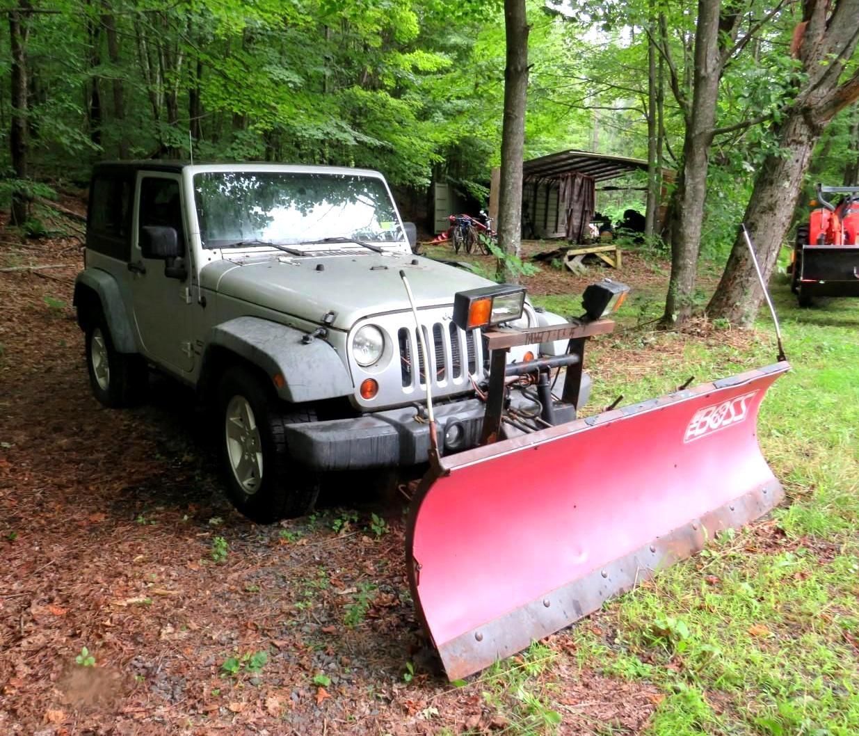 Silver Jeep Wrangler with a red snowplow, parked in a wooded area.