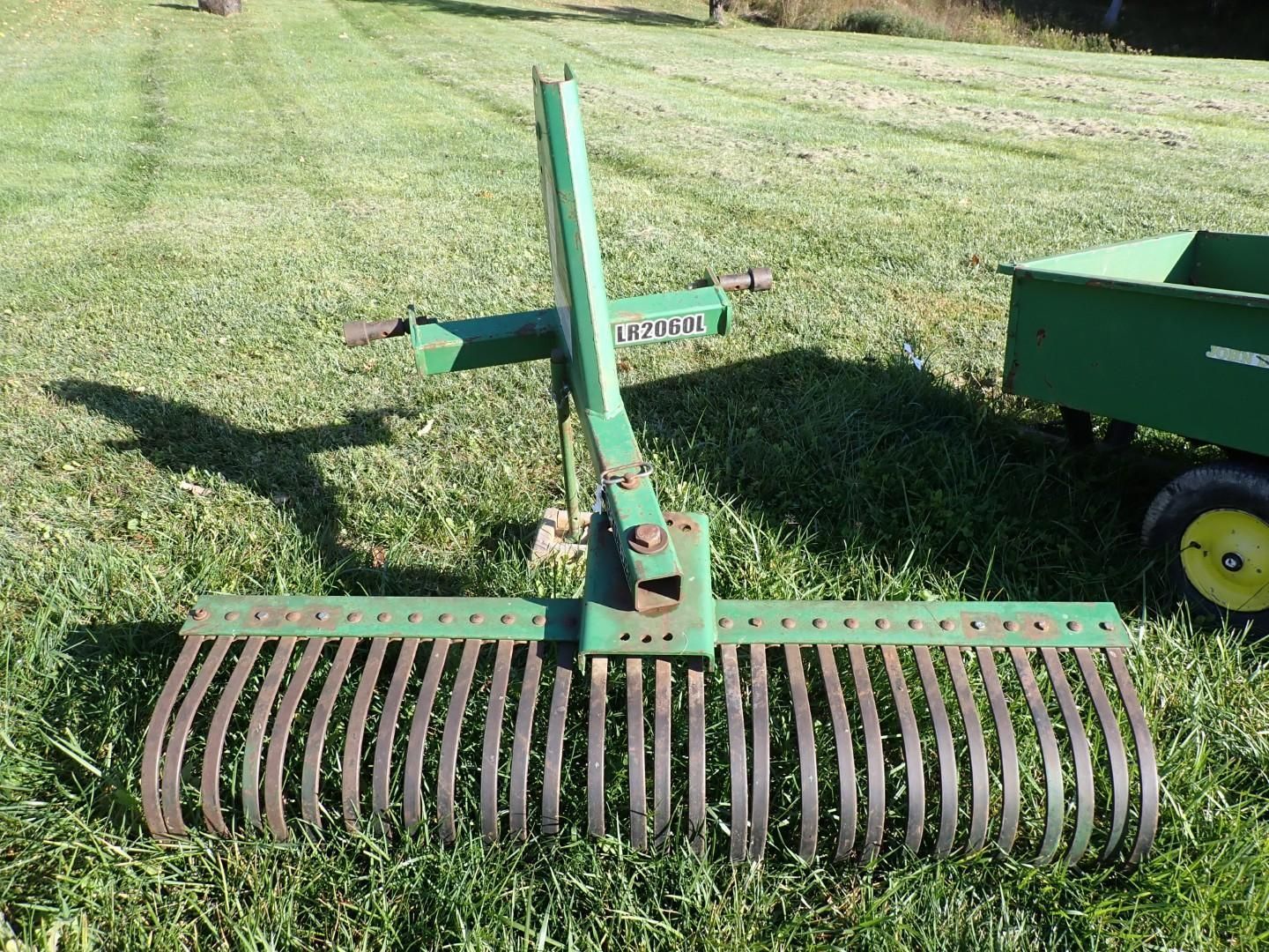 Green landscape rake attachment on grass beside a small trailer.