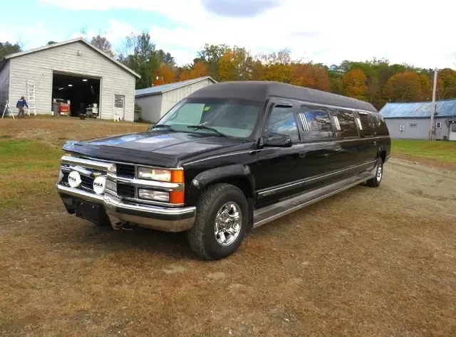 Black limousine parked on brown grass in front of a white building with trees in the background.