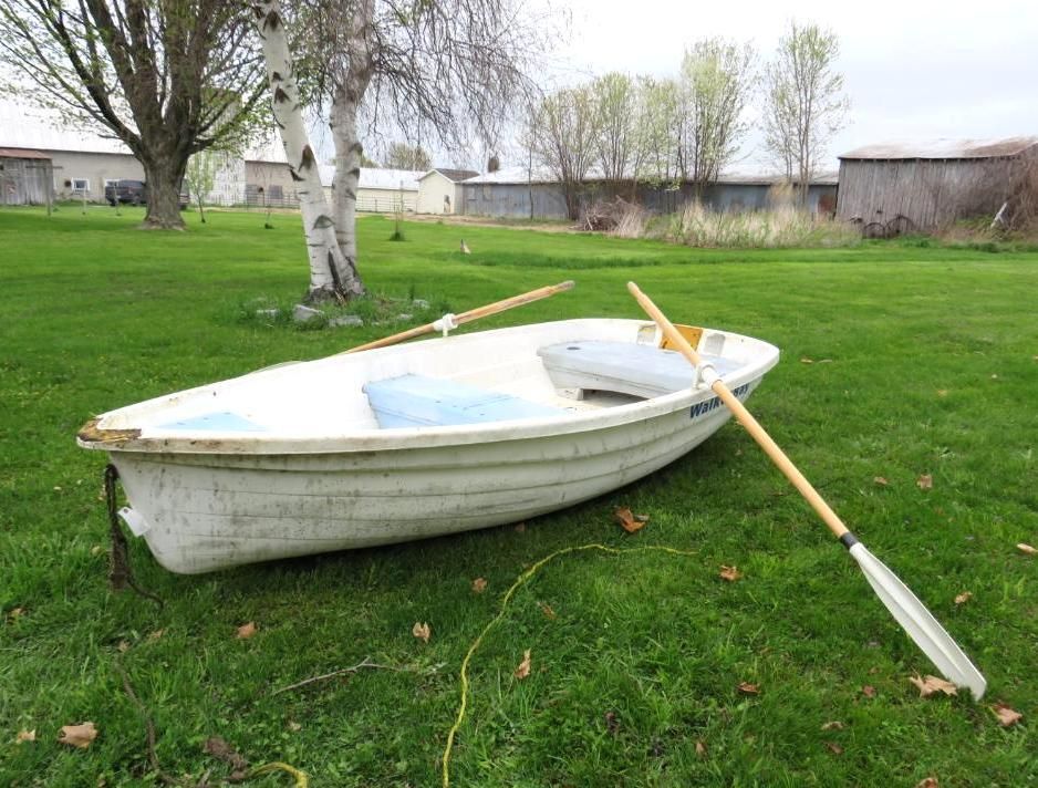 White rowboat on green grass with oars, next to a tree and buildings in the background.