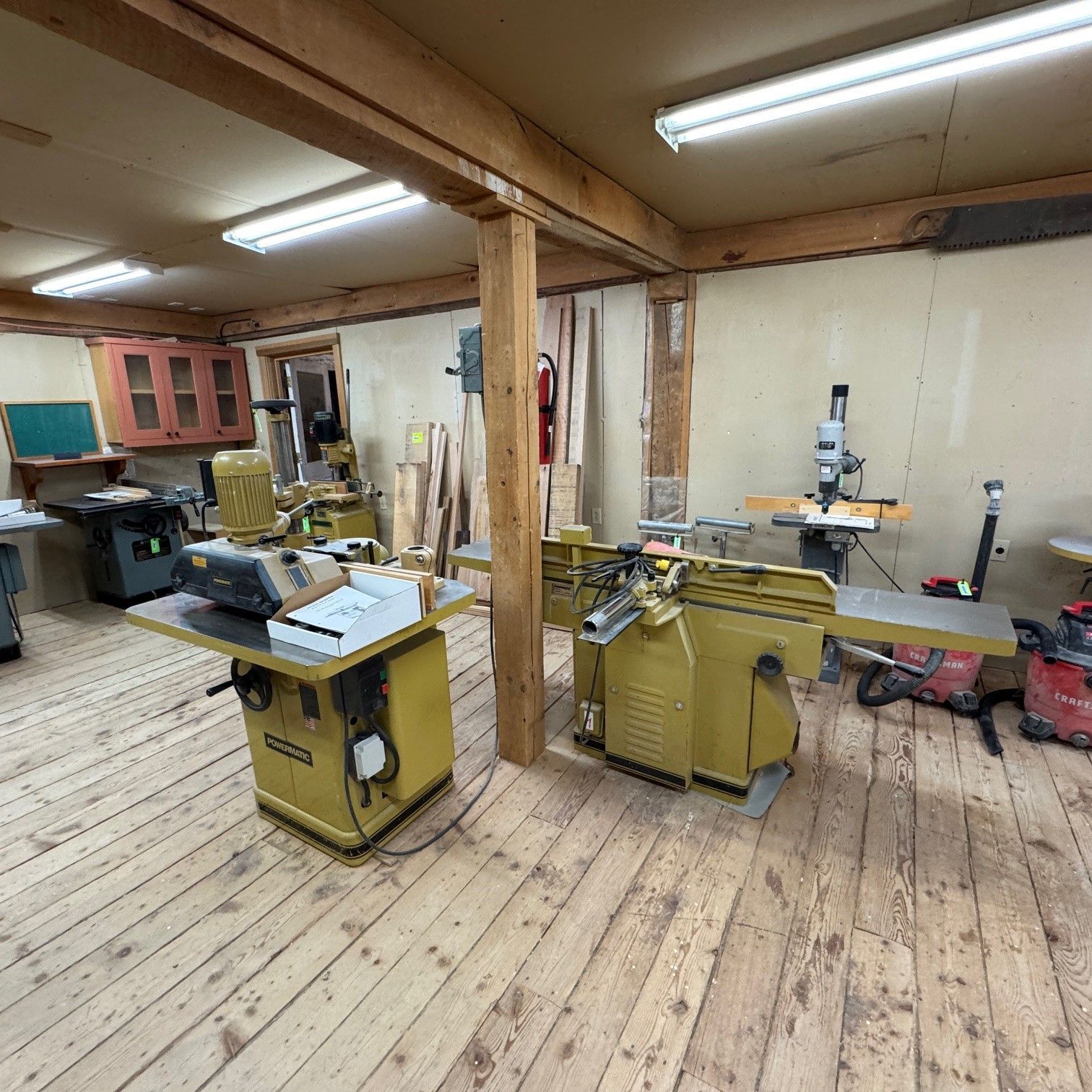Woodshop with two yellow table saws, wooden floor, fluorescent lights, and exposed beams