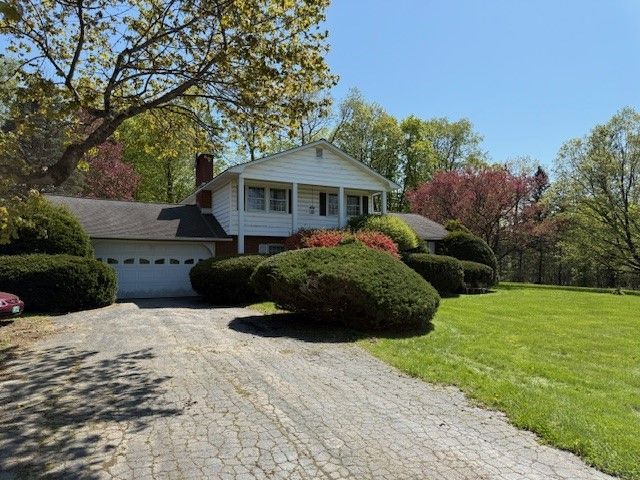 White house with a garage and driveway, surrounded by green grass and trees under a blue sky.