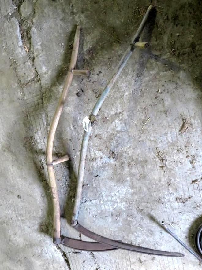 Two antique scythes with curved wooden handles and long blades resting on a concrete surface.