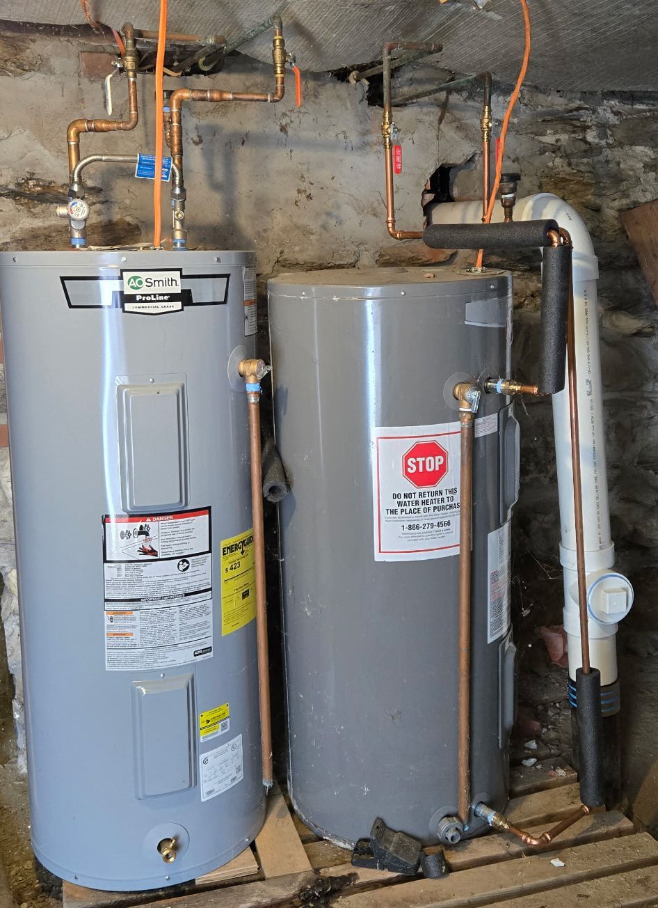 Two grey residential water heaters installed side-by-side in a basement with exposed copper piping and white PVC venting.
