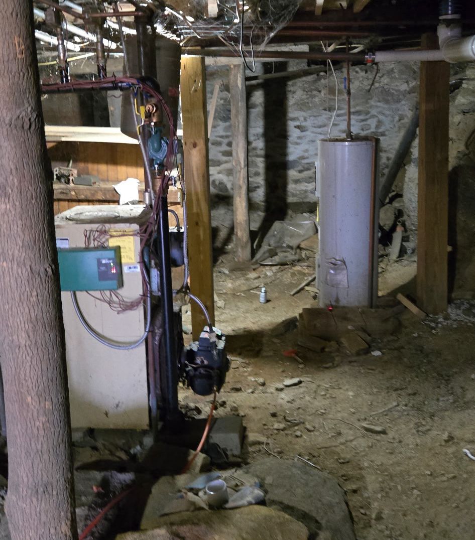 A basement with a dirt floor, stone walls, exposed wooden support beams, a water heater, and a furnace..