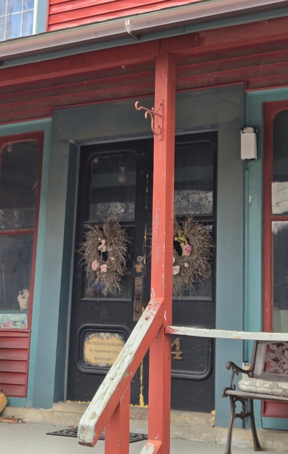 A black double door with two wreaths hangs under a red porch roof, next to a small wooden bench.