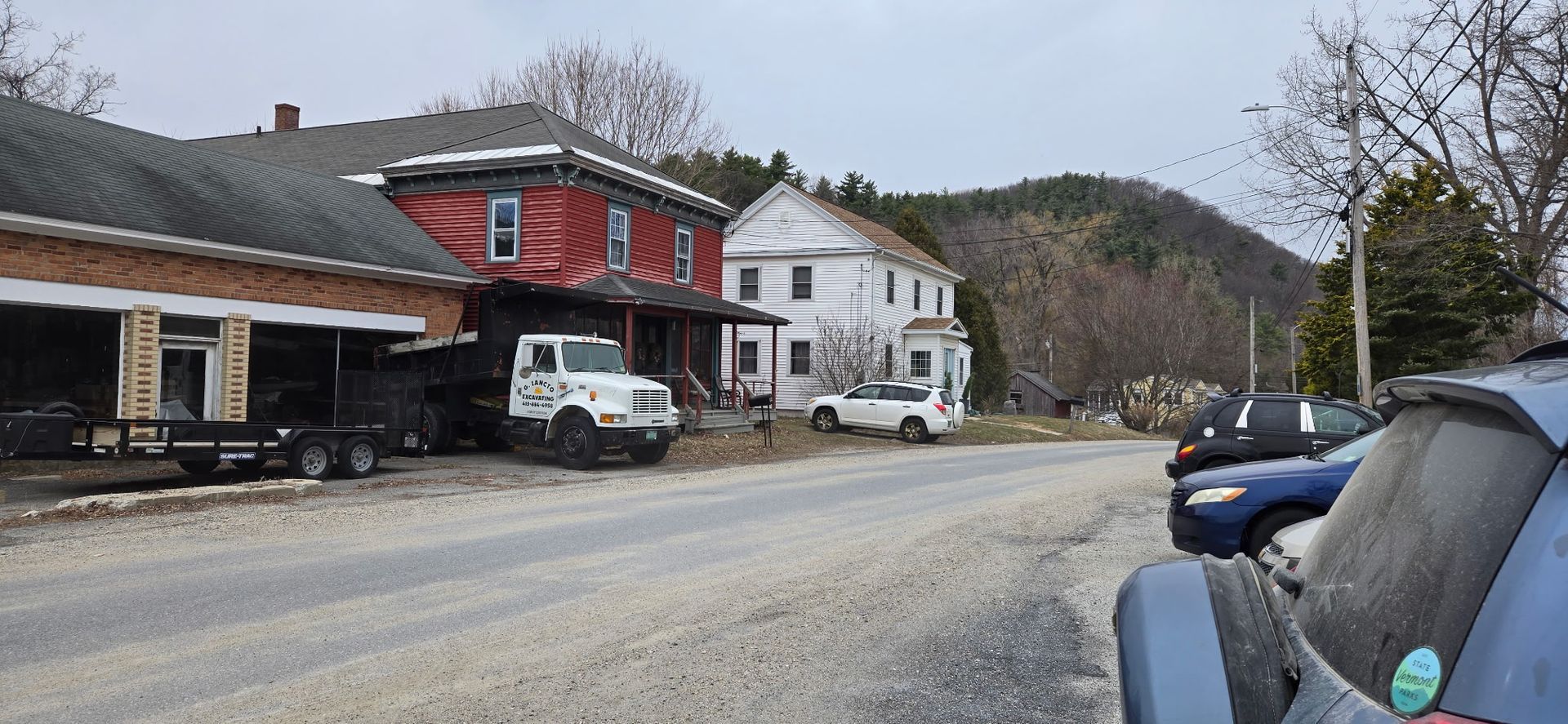 A street scene showing a red building with a white truck, a white house, and parked cars along a gravel shoulder.