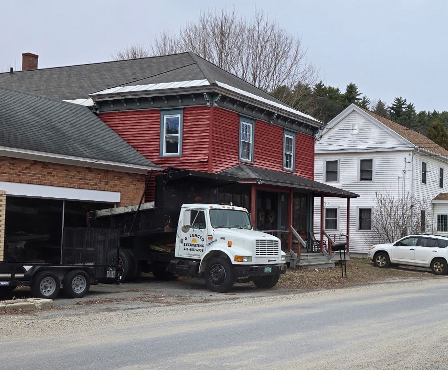 a red, two-story building next to a white house on a gravel roadside.