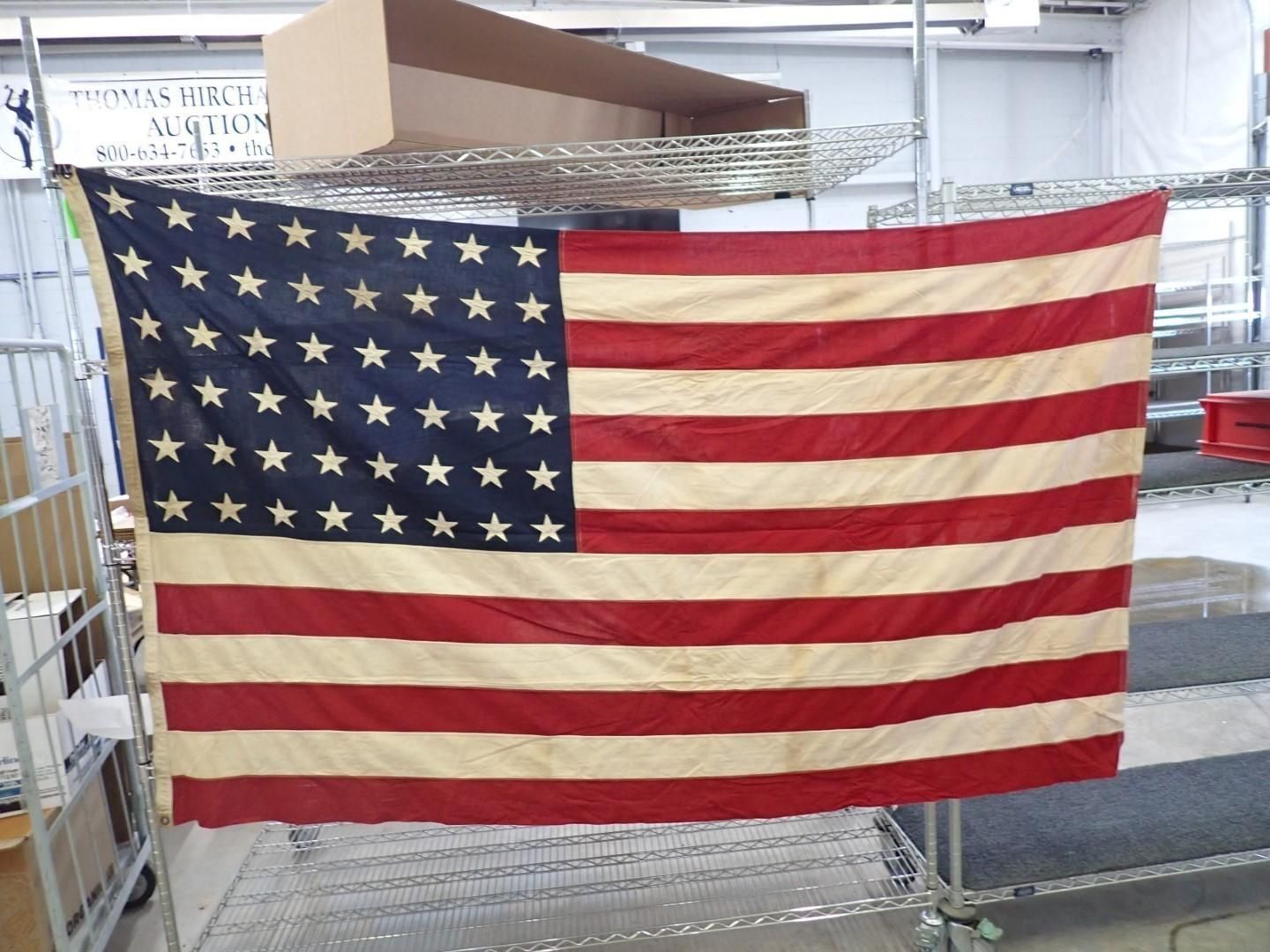 48-star American flag, faded with age, hanging on a metal rack in an industrial setting.