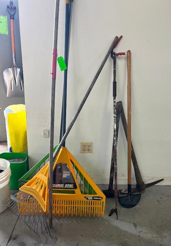 Garden tools, including rakes, shovels, and hoes, lean against a white wall in a garage.