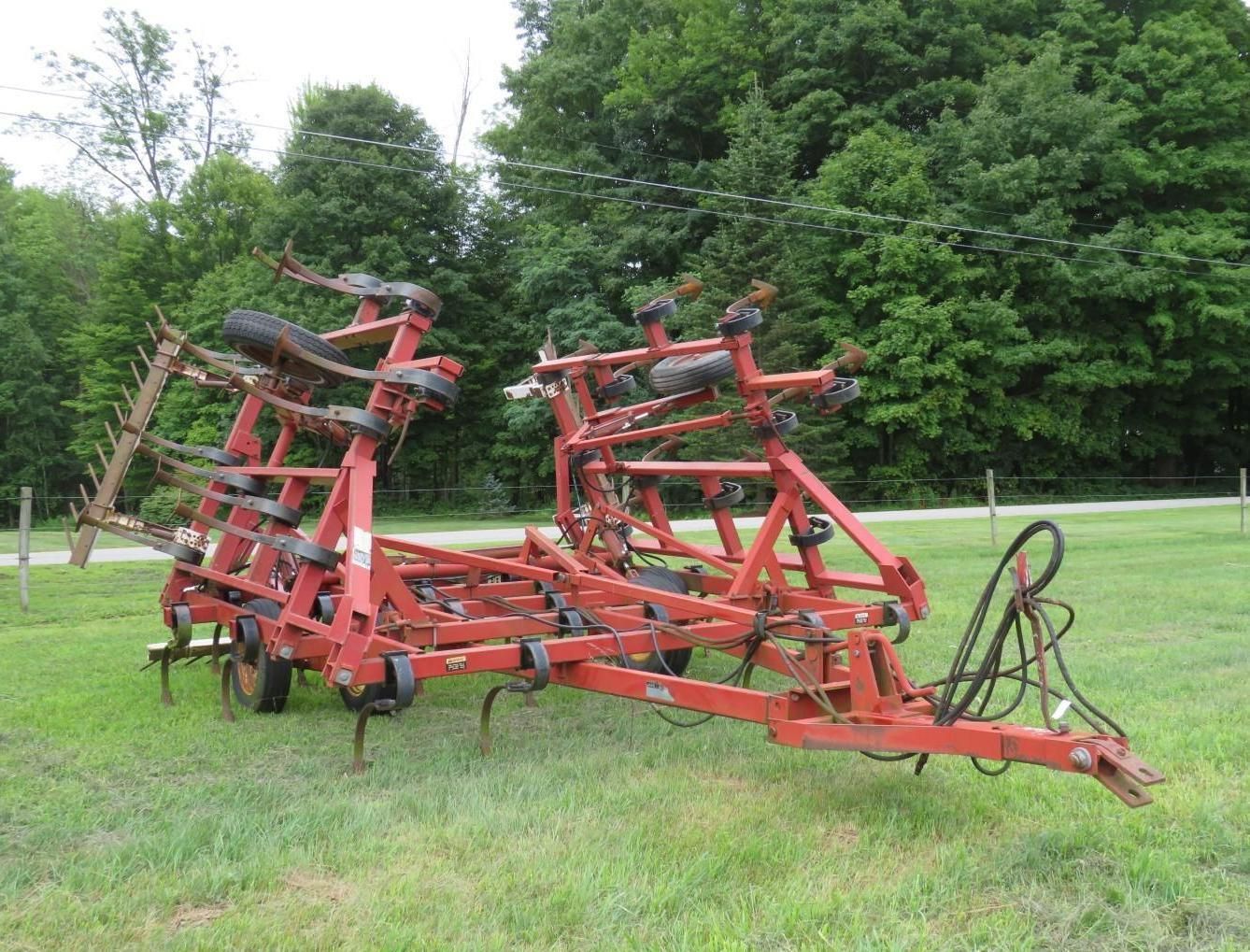 Red agricultural field cultivator on grassy field, near trees.