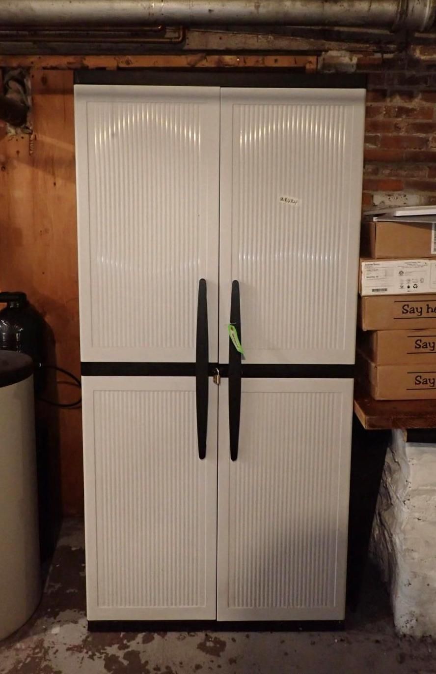 White plastic storage cabinet with black handles and horizontal strap, set against brick wall.