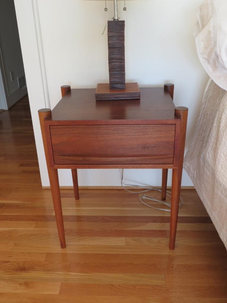 Wooden nightstand with lamp, drawer, and slender legs on wood floor.