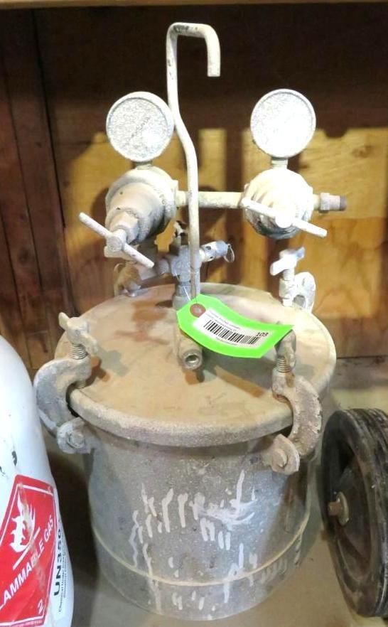 A metal pressure pot with gauges, valves, and a curved handle.