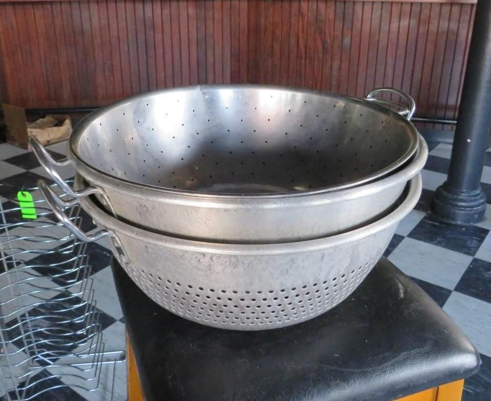 Two metal colanders stacked on a stool, against a checkered floor and wood background.