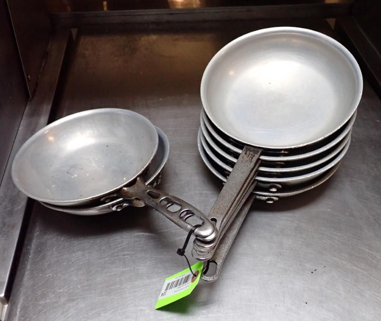 Stack of aluminum frying pans with silver handles, arranged on a stainless steel surface.