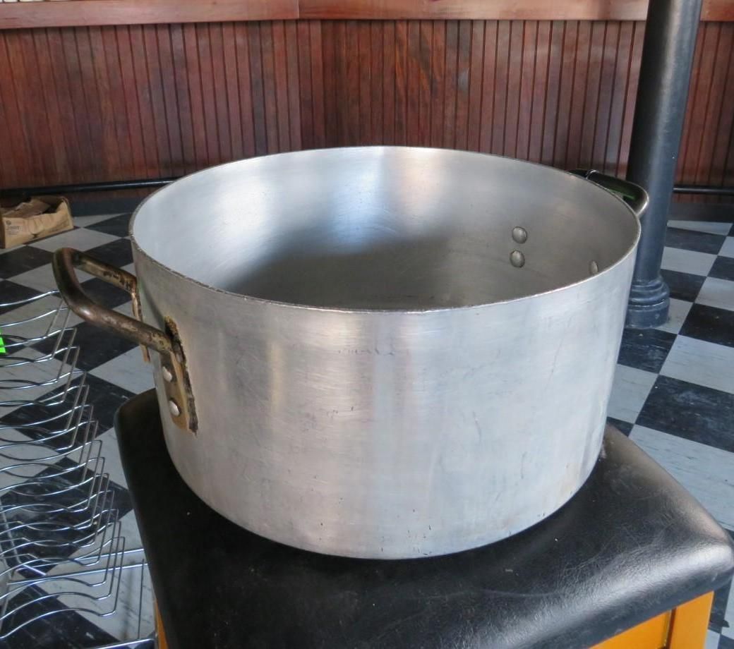 Large, aluminum cooking pot with handles on a black cushion. Indoors, against a wood-paneled wall.