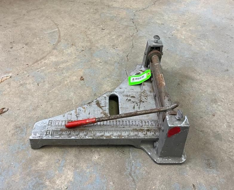 Metal tile cutter on concrete. Gray frame with ruler, red-handled rod, green tag.