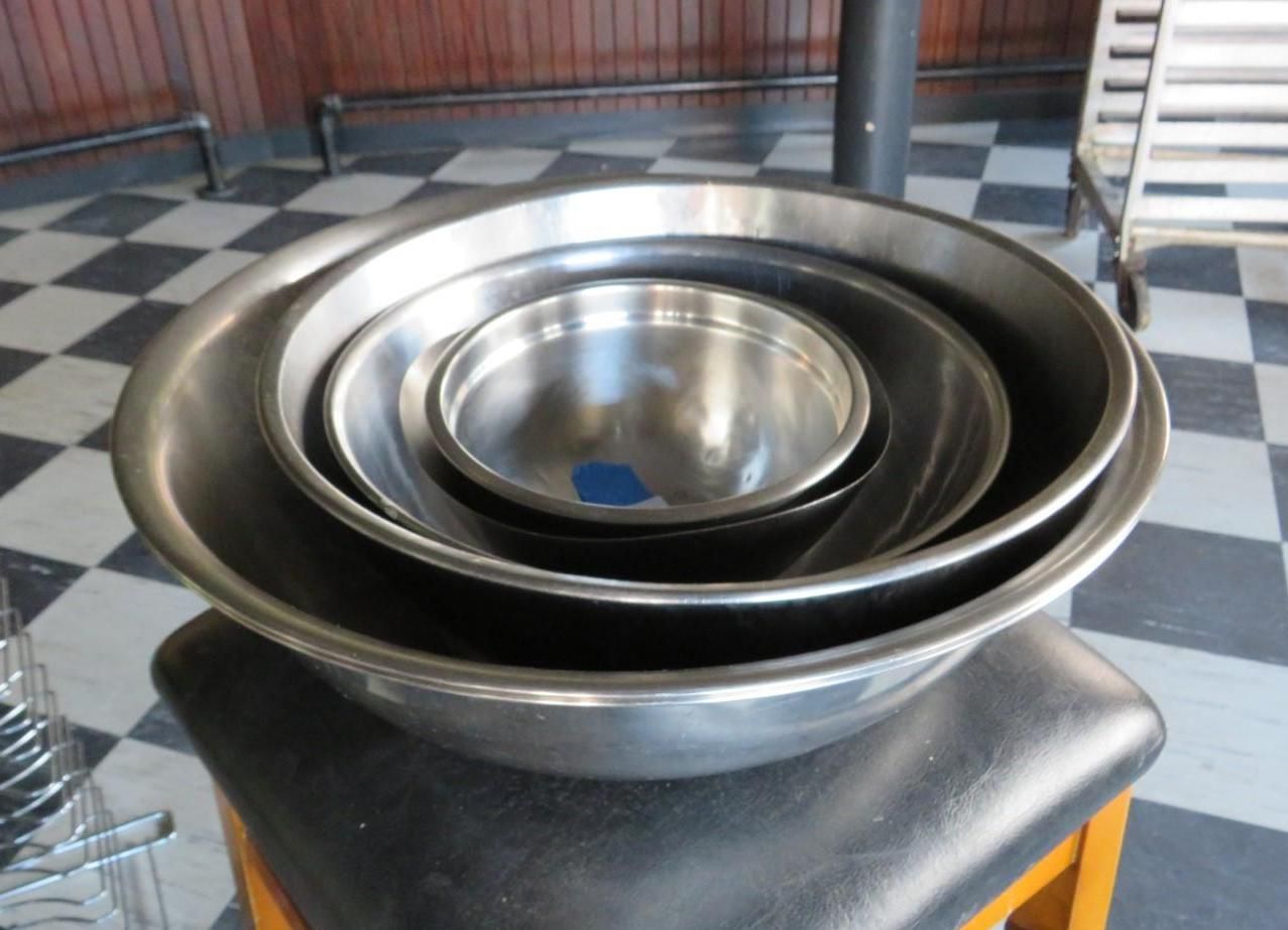 Four stainless steel mixing bowls nested on a black stool, checkered floor in background.