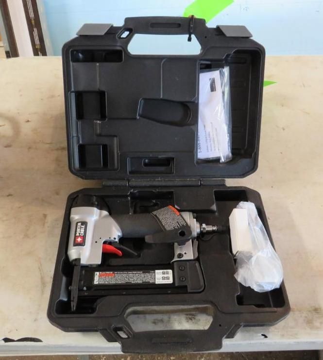 An open black carrying case holds a pneumatic nail gun, accessories, and instruction booklet on a light surface.