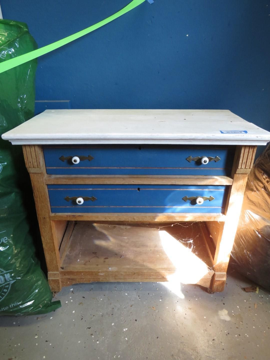 Blue and wood-toned table with white top and two drawers, ornate side supports, and lower shelf.
