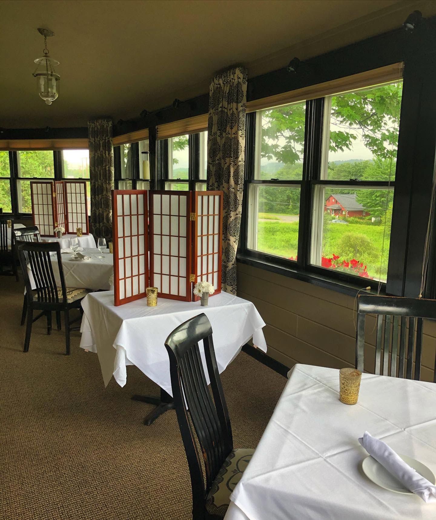 Quiet restaurant dining room with set tables, wicker chairs, and large windows overlooking greenery