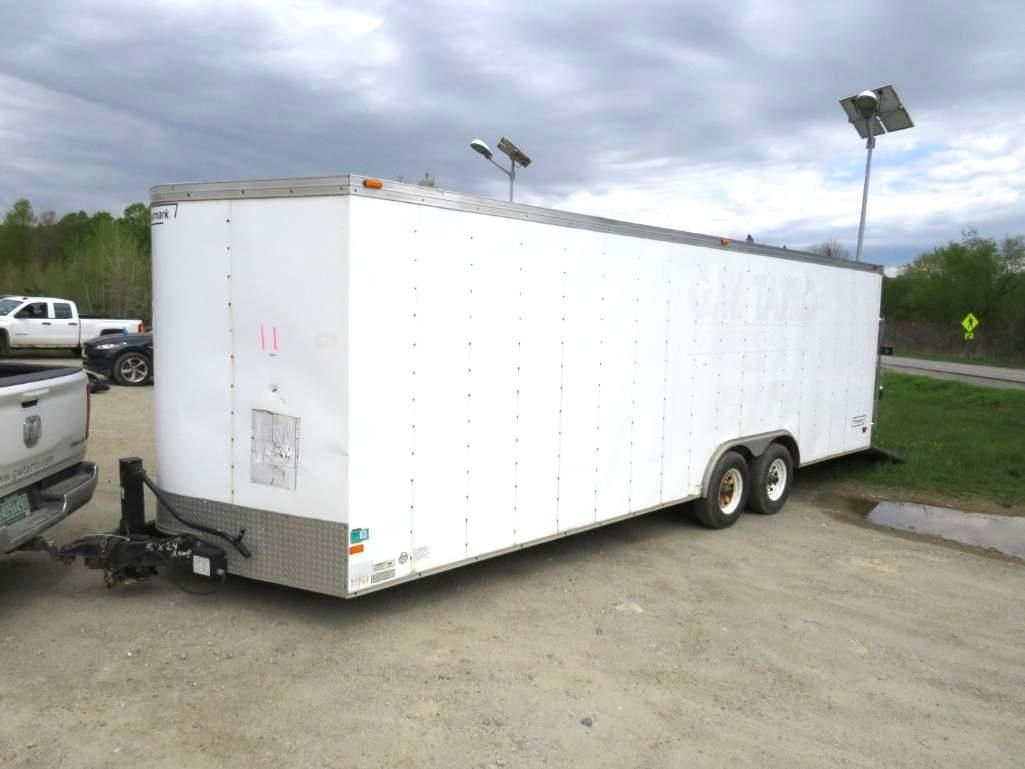 White enclosed trailer hitched to a truck, parked on a paved lot. Cloudy sky in the background.
