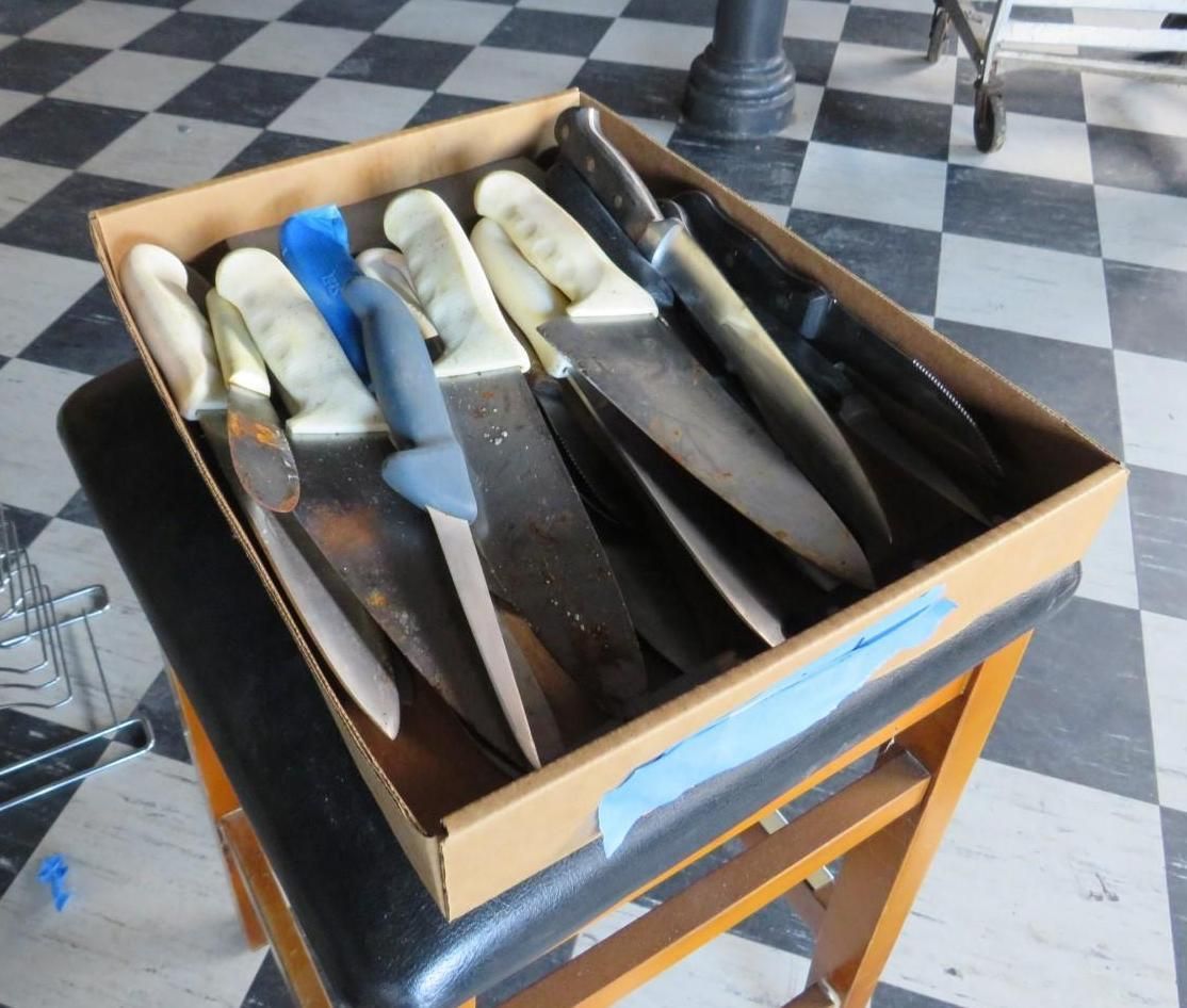 A cardboard box filled with various knives on a small wooden stool with checkered floor in the background.