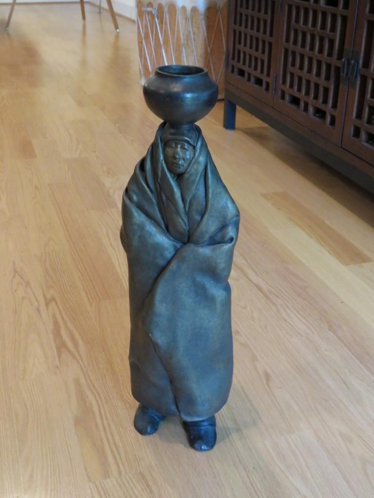 Bronze sculpture of a figure wrapped in a blanket, balancing a pot on its head, standing on a wood floor.
