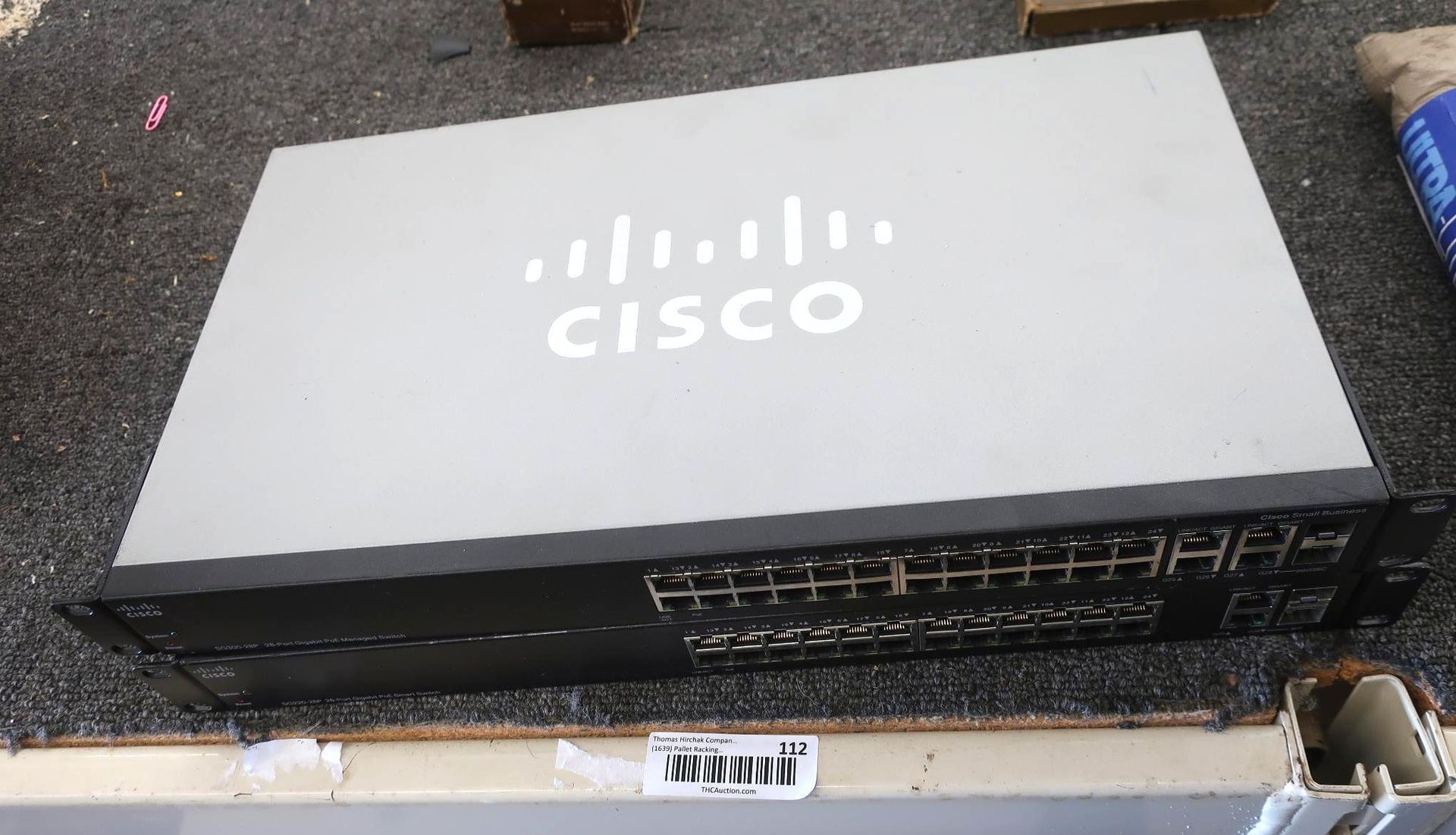 Cisco network switch on a surface. It is gray with a black front panel, containing multiple ports. The Cisco logo is visible.
